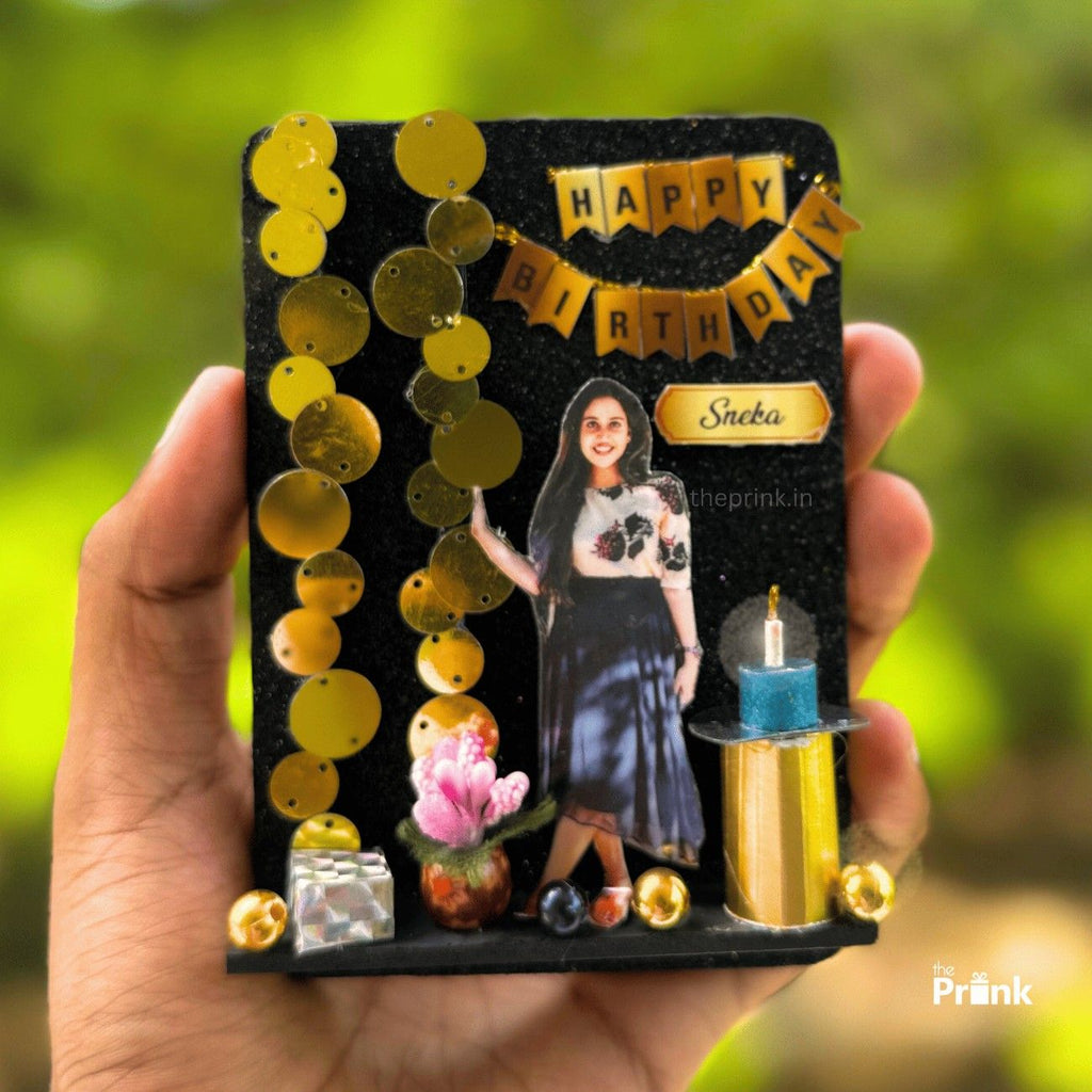 Hand holding a birthday magnet with gold decorations, a female cutout, and a small candle. "Happy Birthday Sneha" text. Green background outdoors.