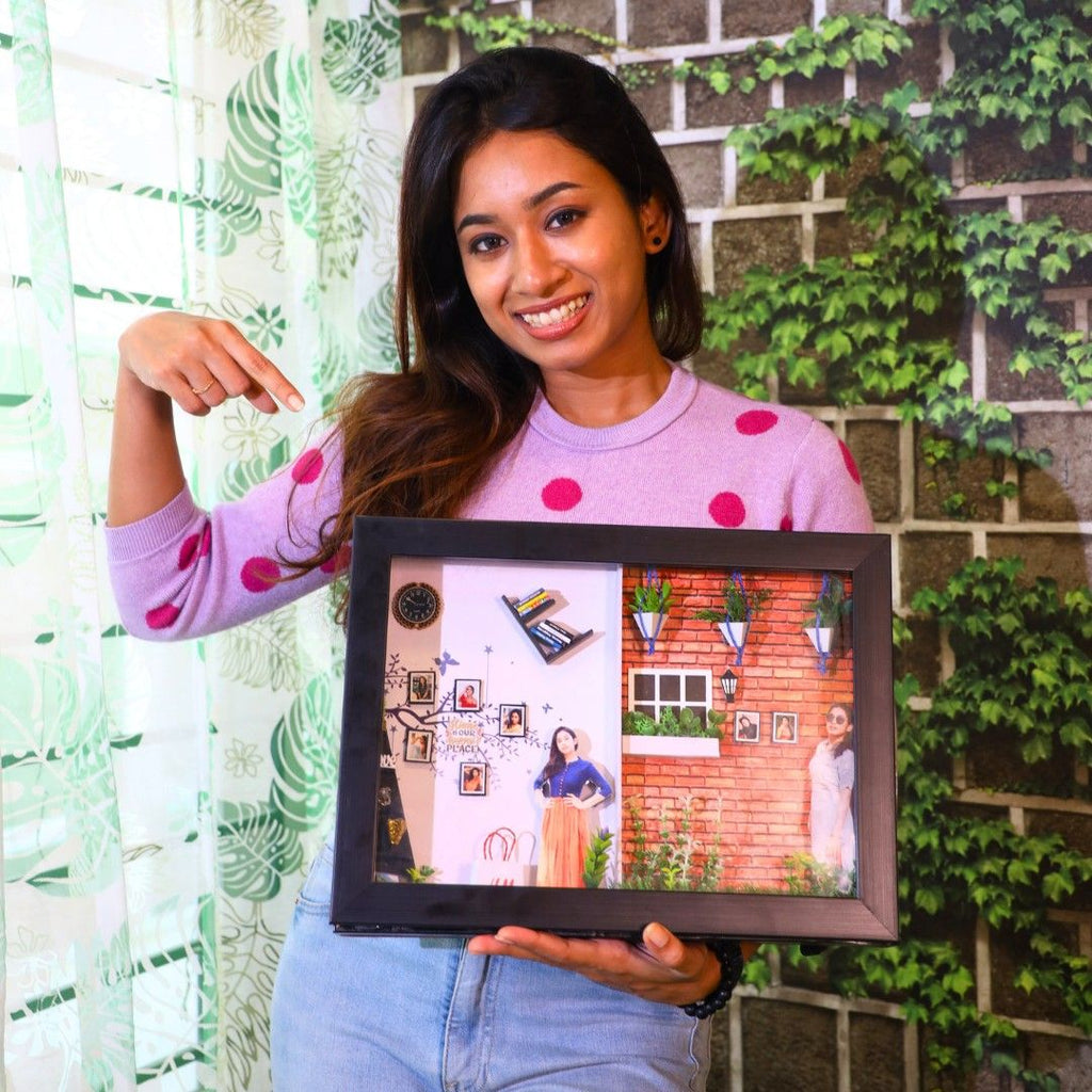 Smiling woman in a sweater holds a framed collage. The background features leafy green vines on a brick wall, creating a cheerful atmosphere.
